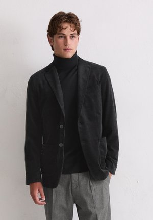 Schwarzer samtiger Blazer mit zwei Knöpfen, mit Reverskragen und Fronttaschen, getragen über einem schwarzen Rollkragenpullover und grauen Hosen.