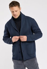 Homme en train de fermer les boutons d'un cardigan en texture bleu marine sur un col roulé noir, portant un pantalon en velours côtelé gris, se tenant contre un fond blanc.