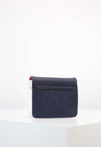 Borsa in pelle sintetica blu navy con patta pieghevole, caratterizzata da lati bianchi e un accento rosso. Texture liscia e forma rettangolare.