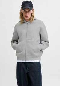 Grå zip-up hoodie lavet af blødt stof med en kengurulomme, kombineret med mørke denim. En sort kasket med et præglogo hviler på hovedet.