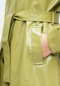 Glanset, lys grønn trenchcoat med belte i midjen. Har en sidelomme og en glatt, teksturert finish. Skreddersydd design med knappedetaljer.