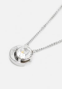 Collana in argento con pendente in cristallo trasparente sfaccettato di grandi dimensioni, racchiuso in una cornice circolare con il nome del marchio "GUESS" inciso.