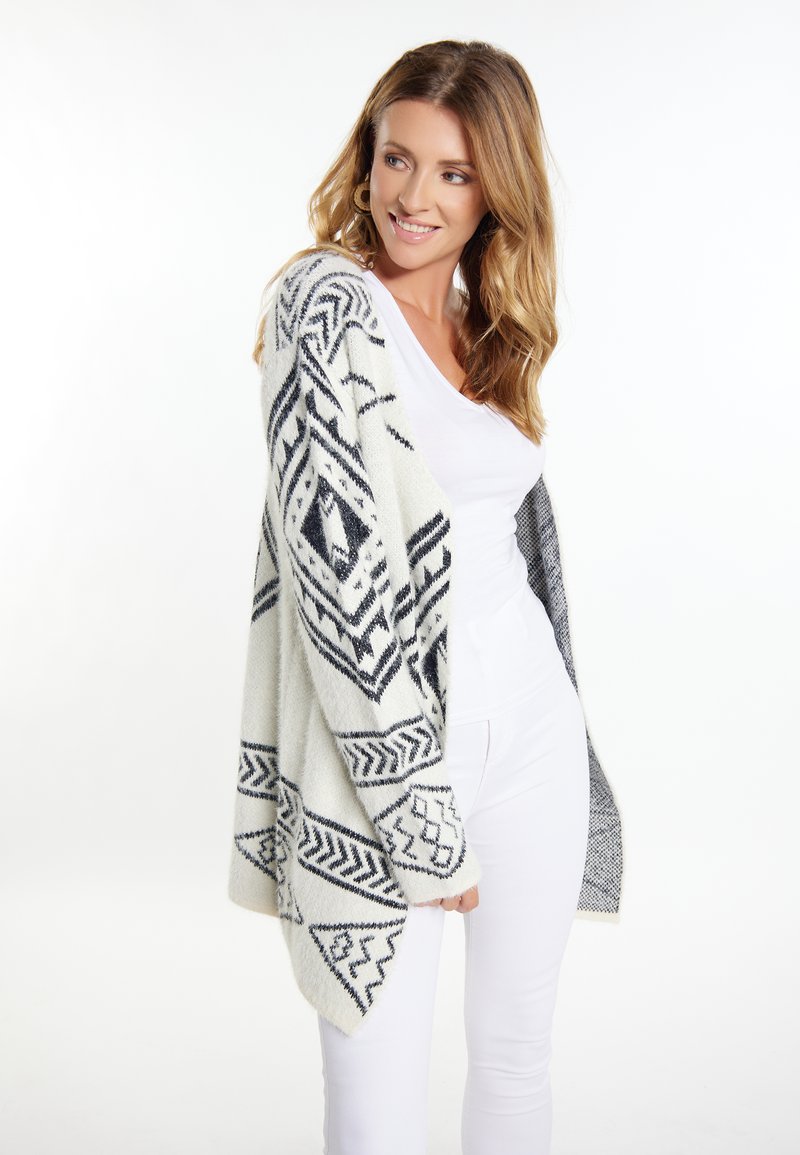Strickjacke in Off-White mit schwarzen geometrischen Mustern, lockerer Schnitt, offene Vorderseite und überschnittene Schultern; weiche Textur, warmes Material.