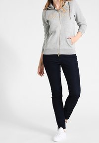 Sudadera gris con cremallera, letras doradas y dos bolsillos laterales, combinada con vaqueros oscuros y zapatillas blancas. Tela suave, mezcla de algodón.