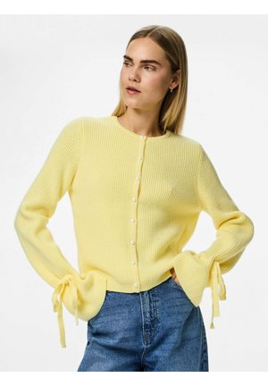 Un cardigan côtelé jaune clair avec des manches longues, des boutons en perles sur le devant et des liens aux poignets. Porté sur un jean en denim bleu.
