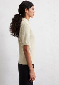 Faalgeel gebreid poloshirt met korte mouwen en een gestructureerd patroon, gecombineerd met een donkere onderkant, weergegeven vanuit een zijprofiel.