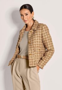 Vrouw poseert in een beige tweed jasje met gouden knopen, een beige top, hoge beige broeken en gouden hoepel oorbellen tegen een effen achtergrond.