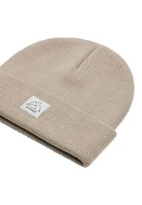 Beige gebreide beanie met een geribbelde textuur en opgerolde manchette, voorzien van een wit logo-label met een geometrisch ontwerp aan de zijkant.