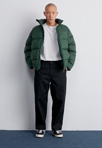 Giacca a vento verde con colletto alto, t-shirt bianca e pantaloni neri. Il modello è in piedi contro un muro semplice, indossando sneakers nere.