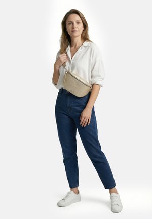 Frau trägt weißes Hemd, blaue Jeans, weiße Turnschuhe und eine beige Umhängetasche, steht vor einem schlichten hellen Hintergrund.