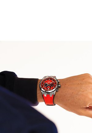Montre de sport rouge avec un boîtier en acier inoxydable, cadran noir et rouge, fonctionnalités chronographe et bracelet en caoutchouc texturé montrant les détails du poignet.