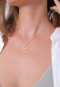 Guldkæde med et hjertemedaljon med en klar �ædelsten som omrids. Fin kæde, minimalistisk design, sat mod en neutral bluse.
