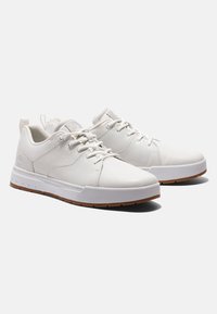 Baskets blanches à tige en cuir lisse, bout arrondi et fermeture à lacets. Elles présentent une semelle en caoutchouc texturée avec un accent marron.