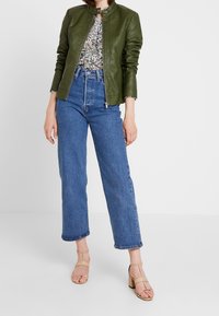 Giacca in pelle verde con collo alto, camicetta floreale, jeans blu a vita alta e sandali con tacco beige; texture liscia, vestibilità casual.