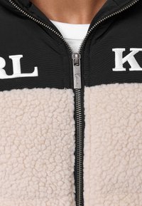 Giacca in pile con zip, caratterizzata da una parte superiore nera con "KARL" in bianco e una parte inferiore beige chiaro testurizzata. Dettaglio della zip in argento.