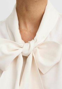 Blusa bianca con un grande fiocco al colletto, realizzata in tessuto morbido, con una texture liscia e una caduta fluida attorno al collo.