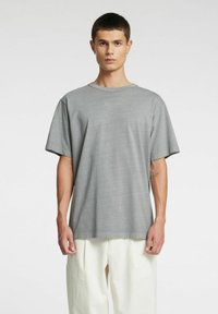 Szara koszulka z krótkim rękawem i okrągłym dekoltem, wykonana z miękkiego materiału. Charakteryzuje się luźnym krojem i subtelną fakturą. Noszona z jasnymi spodniami.