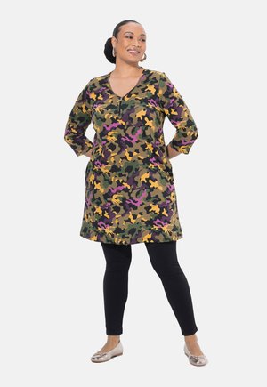 Tunique camouflage verte et violette avec des manches trois-quarts et une fermeture éclair à l'avant. Associée à des leggings noirs et des ballerines argentées.