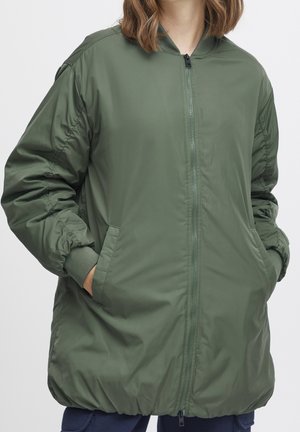 Person wearing a long olive green bomber jacket with hands in side pockets, standing against a plain light background.