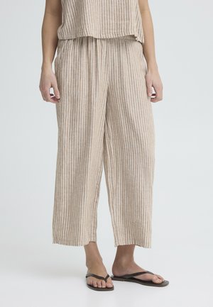 Kvinde iført beige og hvide bukser med lodrette striber og sorte flip-flops, stående mod en ensfarvet baggrund.