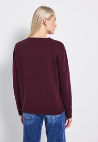 Pull bordeaux à coupe décontractée, avec un col et des poignets côtelés. Porté avec un jean bleu. Le tissu semble doux et texturé.