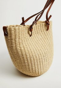 Sac tote en paille tissée avec une forme arrondie, de couleur beige naturel, et des poignées en cuir marron fixées avec un matériel en métal.