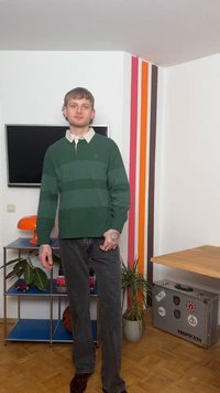 Νεαρός άνδρας με τατουαρισμένα χέρια, φορώντας πράσινο ριγέ πουλόβερ και σκούρο τζιν, στέκεται σε ένα δωμάτιο με πολύχρωμες κάθετες ρίγες σε λευκούς τοίχους.