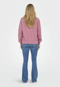 Felpa rosa con polsini e fondo a coste, con dettagli a occhio sulle maniche. Abbinata a jeans a zampa d'elefante blu e sneakers bianche.