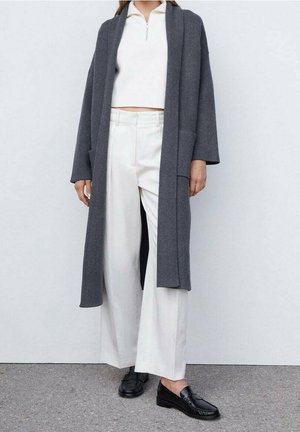Person trägt einen dunkelgrauen langen offenen Cardigan, einen weißen kurzen Reißverschluss-Pullover, weiße weit geschnittene Hosen und schwarze Slip-On-Schuhe und steht vor einer hellen Wand.