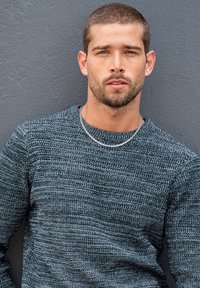Grau strukturierter Pullover mit Rundhalsausschnitt und geripptem Muster; kombiniert mit einer Silberkette. Das Model hat kurze Haare und einen Bart.