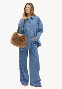 Mujer con una camisa vaquera azul de gran tamaño y vaqueros de pierna ancha con lentejuelas multicolores, sosteniendo un gran bolso marrón difuso de hombro.