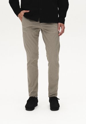 Uomo che indossa pantaloni beige slim fit, scarpe nere e camicia nera abbottonata con una mano in tasca su sfondo bianco.