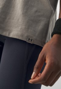 Gros plan d'une personne portant un t-shirt gris Under Armour, des leggings noirs et un bandeau noir au poignet, main détendue sur le côté.