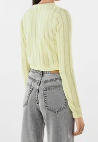 Kobieta w jasnożółtym, długim sweterku z dzianiny i wysokich, szarych dżinsach z tyłu na gładkim tle.