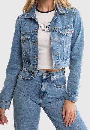 Chaqueta de mezclilla azul claro cortada con cierre de botones, bolsillos en el pecho y una textura ligeramente desgastada, combinada con una camiseta gráfica blanca y jeans.