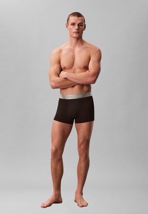 Muskularny mężczyzna stojący boso z założonymi na piersiach ramionami, ubrany w czarne bokserki Calvin Klein na jednolitym szarym tle.