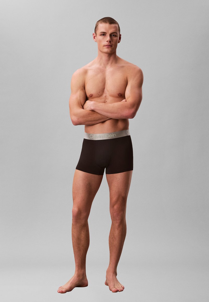 Homme musclé debout pieds nus, les bras croisés, portant un boxer Calvin Klein noir sur un fond gris uni.