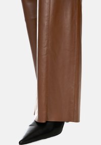 Brown leather trousers with a smooth texture and straight cut, paired with pointed black heeled shoes. Visible seam details enhance the design.