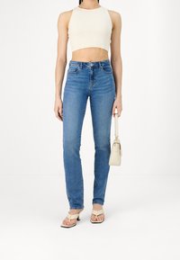 Jeans de tiro alto de color azul claro, pierna ajustada, lavado desvaído, combinados con un top corto color crema y sandalias de tacón blancas, sosteniendo un pequeño bolso beige.