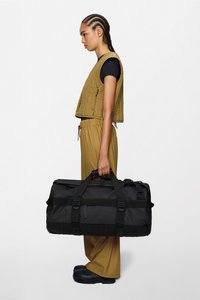 Sac polochon noir avec poignées doubles, large ouverture et sangles latérales. Modèle portant une veste matelassée olive et un pantalon assorti, debout horizontalement.