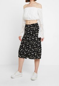 Valkoinen olkaimeton lyhyt paita, jossa on elastinen kaula-aukko ja pitsi-hihat, yhdistetty mustan kukallisen midi-hameen kanssa, joka on käytössä valkoisten tennareiden kanssa.