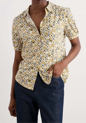 Kobieta w koszuli na krótki rękaw zapinanej na guziki z geometrycznym kwiatowym wzorem w kolorach niebieskim, białym i musztardowym, w połączeniu z ciemnoniebieskimi dżinsami.