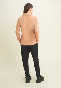 Donna con capelli castani lisci, che indossa una maglietta beige a maniche lunghe e pantaloni neri, in piedi con le mani in tasca, rivolta di spalle.