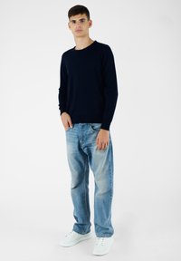 Pullover girocollo navy abbinato a jeans chiari strappati e sneakers bianche, con vestibilità comoda e hardware minimale.