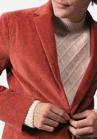 Roestkleurige corduroy blazer met ontplooide revers, voorzakken en een soepele afwerking, gelaagd over een crèmekleurig, gestructureerd sweater.