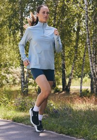 Lys blå langermet treningstopp med glidelås i kragen, kombinert med mørke shorts og svarte løpetøfler, plassert på en skogkledd utendørs sti.