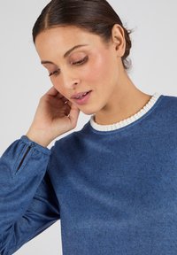 Blauwe gebreide top met een textuurafwerking, voorzien van een golvende witte kraag en knoopdetail bij de manchetten van de mouwen.