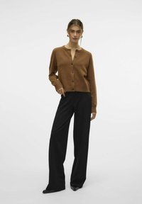 Hnědý, zkrácený cardigan s kulatým výstřihem a zapínáním na knoflíky, v kombinaci s černými kalhotami s širokými nohavicemi. Hladká textura, minimalistický design.