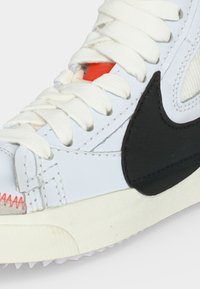 Close-up of white sneaker with thick white laces, black Nike swoosh, red stitch detail, and a hint of orange under the laces.