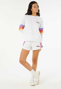 Sweatshirt en coton blanc avec des manches rayées multicolores. Associé à un short blanc arborant un logo coloré. Baskets blanches avec une texture visible.